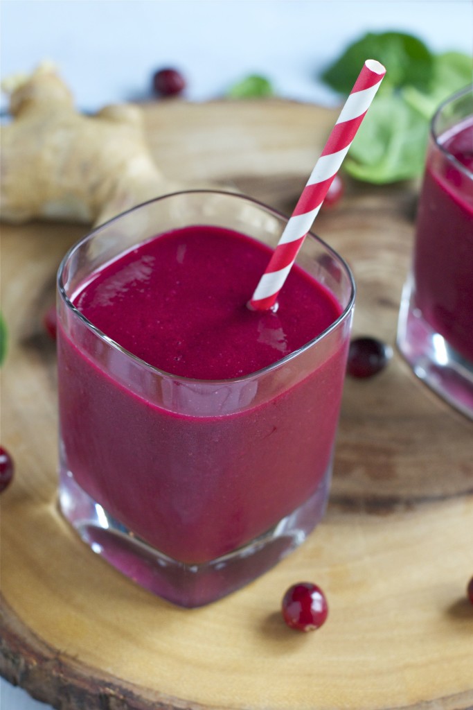 Close-up of a glass filled with vibrant cranberry beet smoothie, topped with a red and white striped straw, set on a wooden board with scattered cranberries and fresh ginger in the background.