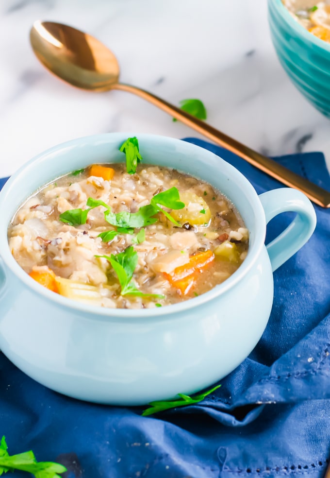 A light blue bowl filled with creamy Crockpot Chicken and Rice Soup, topped with fresh parsley. Chunks of carrots and celery are visible in the soup. The bowl sits on a blue cloth with a gold spoon in the background.