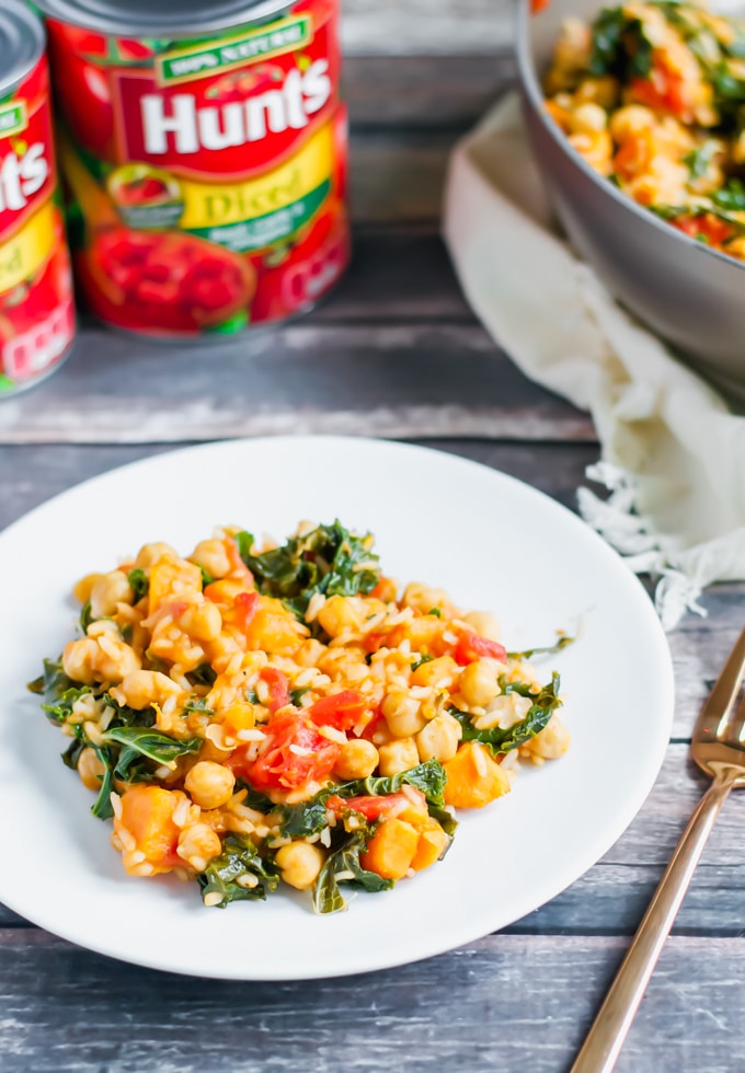 A white plate with a serving of chickpea, tomato, and kale stew sits on a wooden table. Nearby are cans of Hunt’s diced tomatoes and a bowl of more One Pot Sweet Potato Skillet stew. A gold fork rests next to the plate.