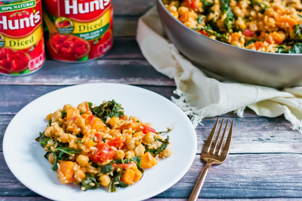 A white plate with a serving of vegetable stew—reminiscent of a One Pot Sweet Potato Skillet—featuring chickpeas, tomatoes, and greens sits next to a gold fork. Behind it are cans of diced tomatoes and a pan with more stew on a wooden table.