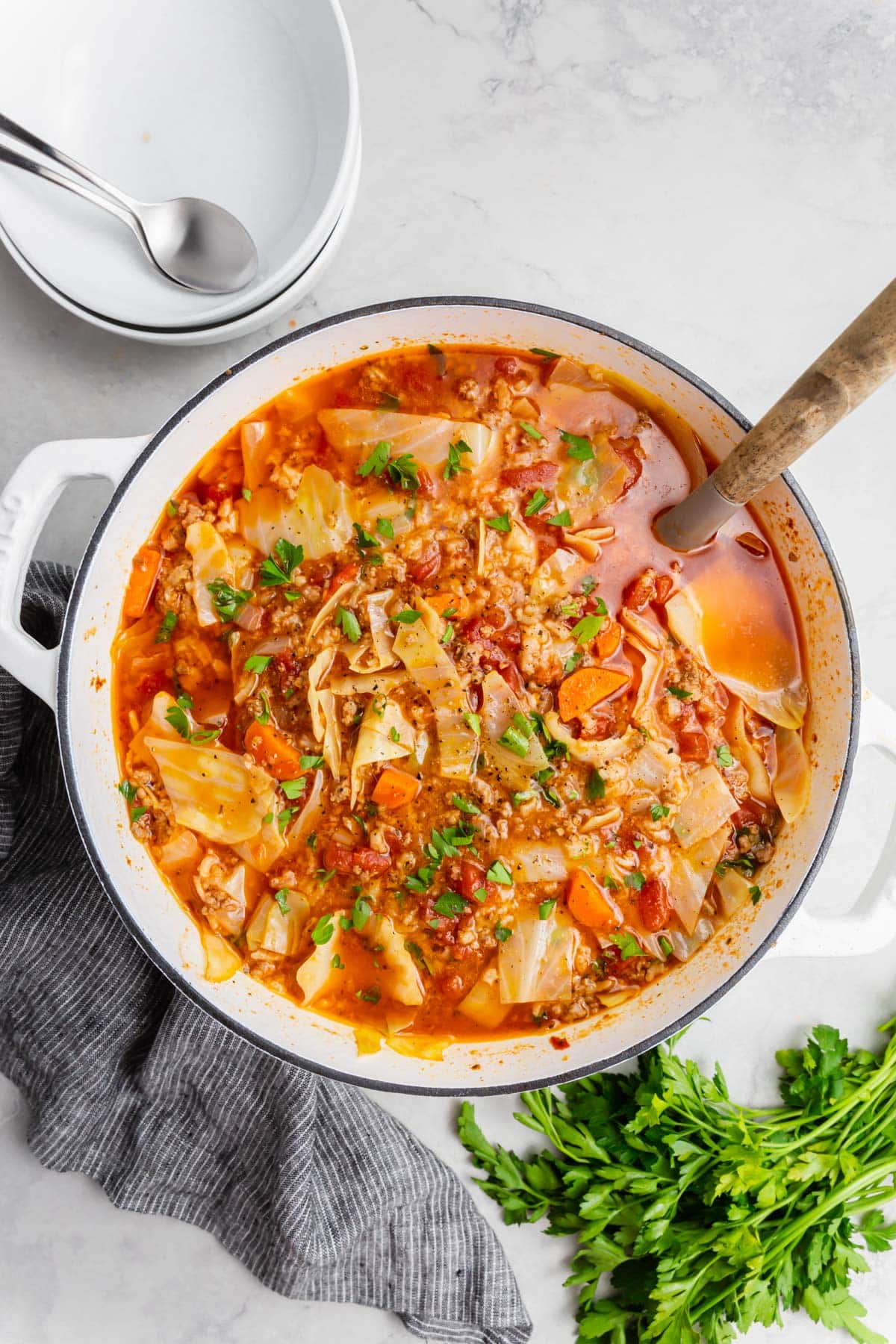 An overhead view of a dutch oven of cabbage roll soup with a spoon in it with a bunch of flat leaf parsley on the side and empty bowls with spoons on the side.