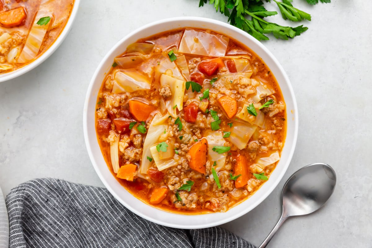 A bowl of Cabbage Roll Soup with ground meat, carrot slices, tomato chunks, and chopped parsley sits on a white surface beside a spoon, a gray striped napkin, and some fresh parsley.