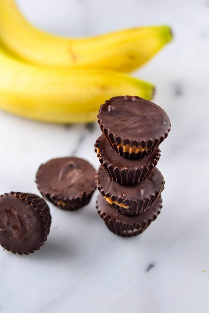 A stack of homemade Chocolate Peanut Butter Banana Bites is arranged on a marble surface, with two bananas in the background and two additional chocolate cups lying nearby.
