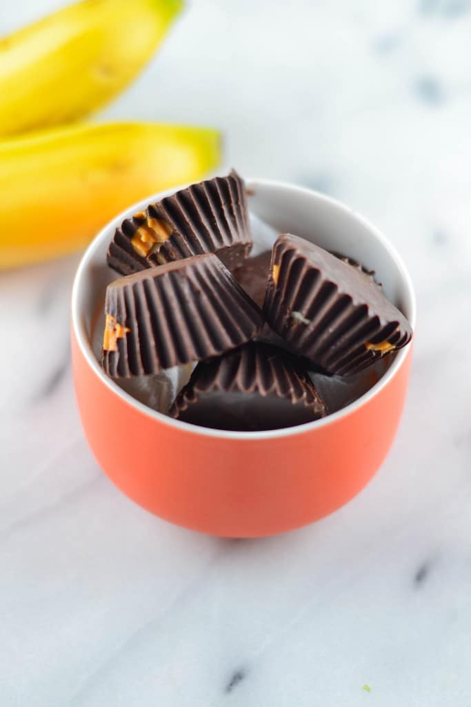 A small orange bowl filled with dark chocolate cups, some with peanut butter filling visible. In the background, two yellow bananas rest on a white marble surface—perfect ingredients for Chocolate Peanut Butter Banana Bites.