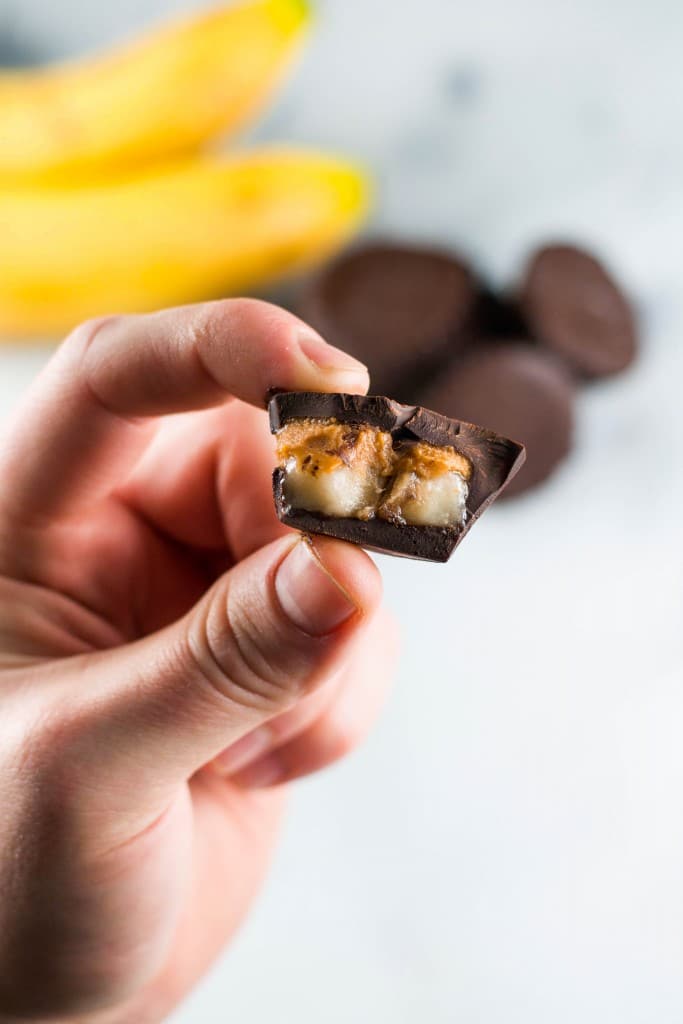 A hand holds a piece of chocolate with a banana filling inside. In the background, blurred bananas and more Chocolate Peanut Butter Banana Bites rest on a light surface.