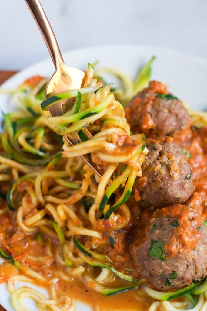 A fork twirls zucchini noodles coated in tomato sauce, next to three turkey meatballs on a white plate. This Zoodles with Turkey Meatballs dish is garnished with fresh herbs and served with extra sauce.