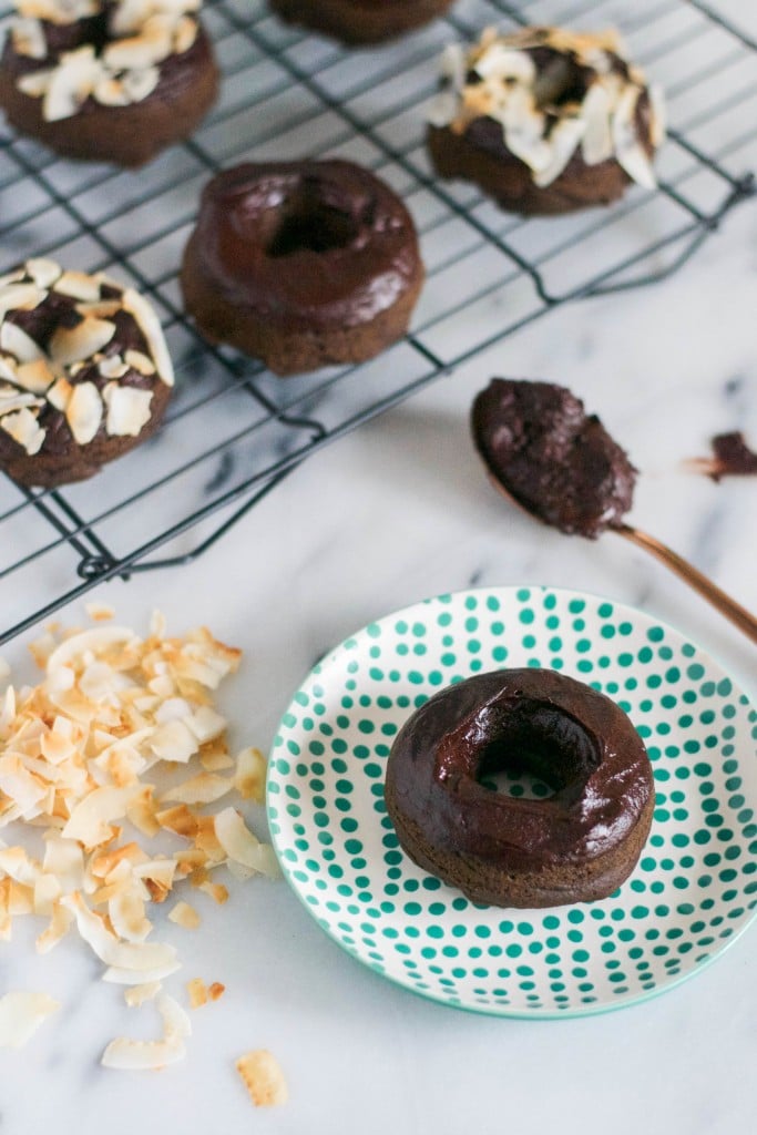 A chocolate donut on a plate in front of a cooling rack with chocolate donuts and a pile of toasted coconut shreds.