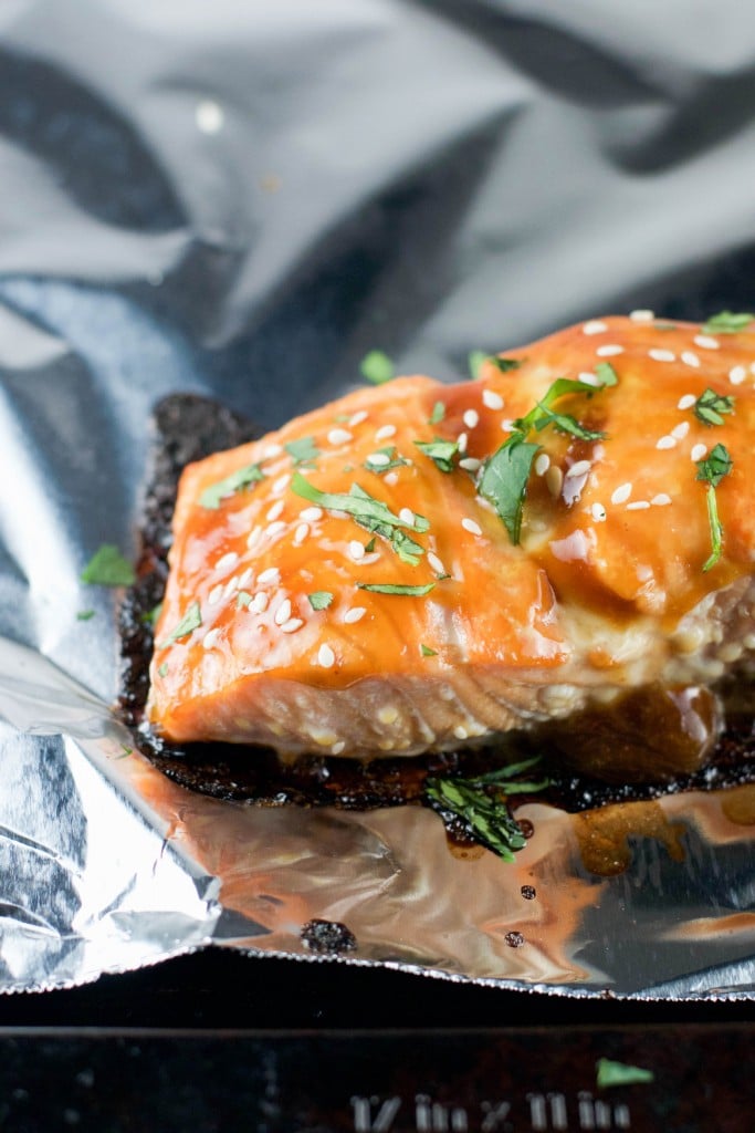A Gluten-Free Baked Teriyaki Salmon fillet is glazed, then topped with sesame seeds and chopped herbs, resting on a sheet of aluminum foil.