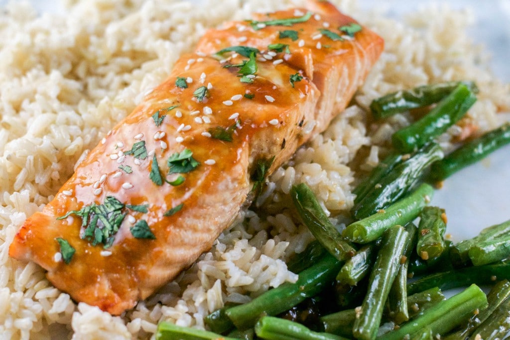 A gluten-free baked teriyaki salmon fillet glazed to perfection, topped with sesame seeds and herbs, is served on a bed of rice with a side of green beans.