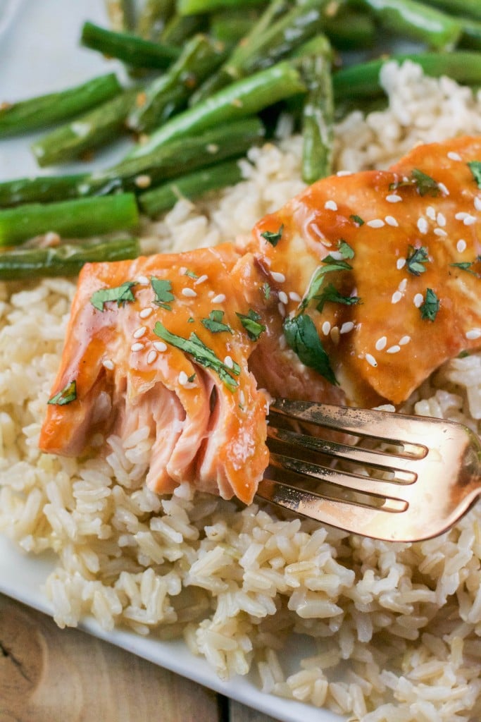 A close-up of a fork flaking a piece of Gluten-Free Baked Teriyaki Salmon, glazed and garnished with sesame seeds and herbs, served on brown rice with green beans in the background.