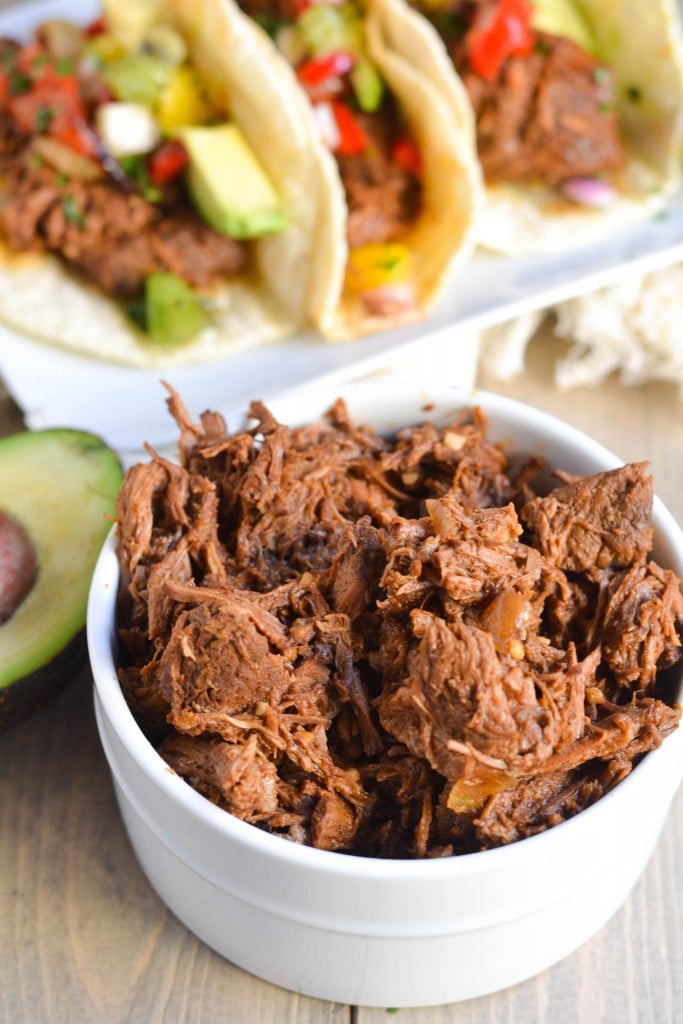 A white bowl filled with Crockpot Shredded Beef sits in the foreground, with soft tacos filled with beef, avocado, and vegetables in the background. A halved avocado rests beside the bowl on a wooden surface.
