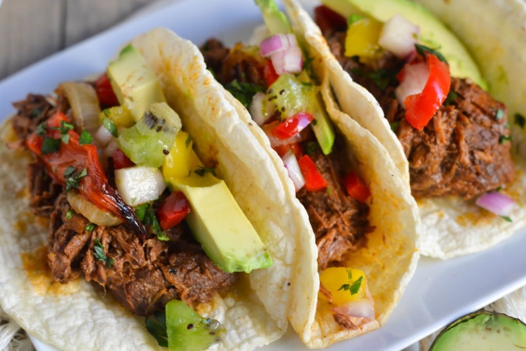 Three soft tortillas filled with Crockpot Shredded Beef Tacos, diced avocado, chopped onions, red and yellow bell peppers, and kiwi pieces, garnished with herbs and served on a white plate.