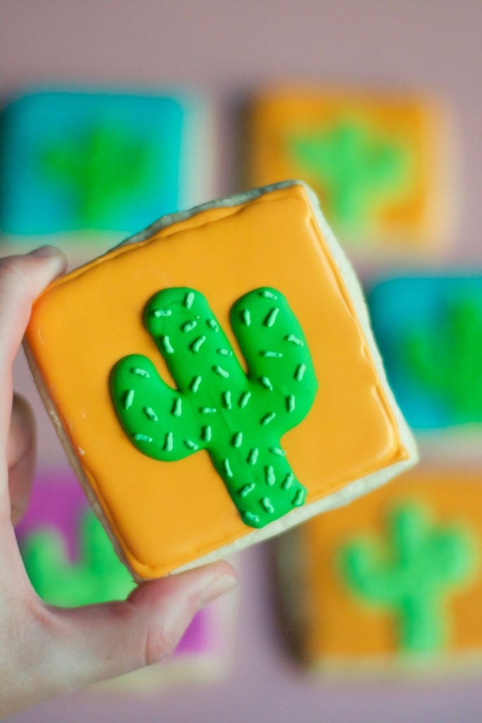 A hand holds a gluten-free cactus sugar cookie decorated with orange icing, a green cactus design, and white sprinkles, with similar colorful cactus cookies blurred in the background.