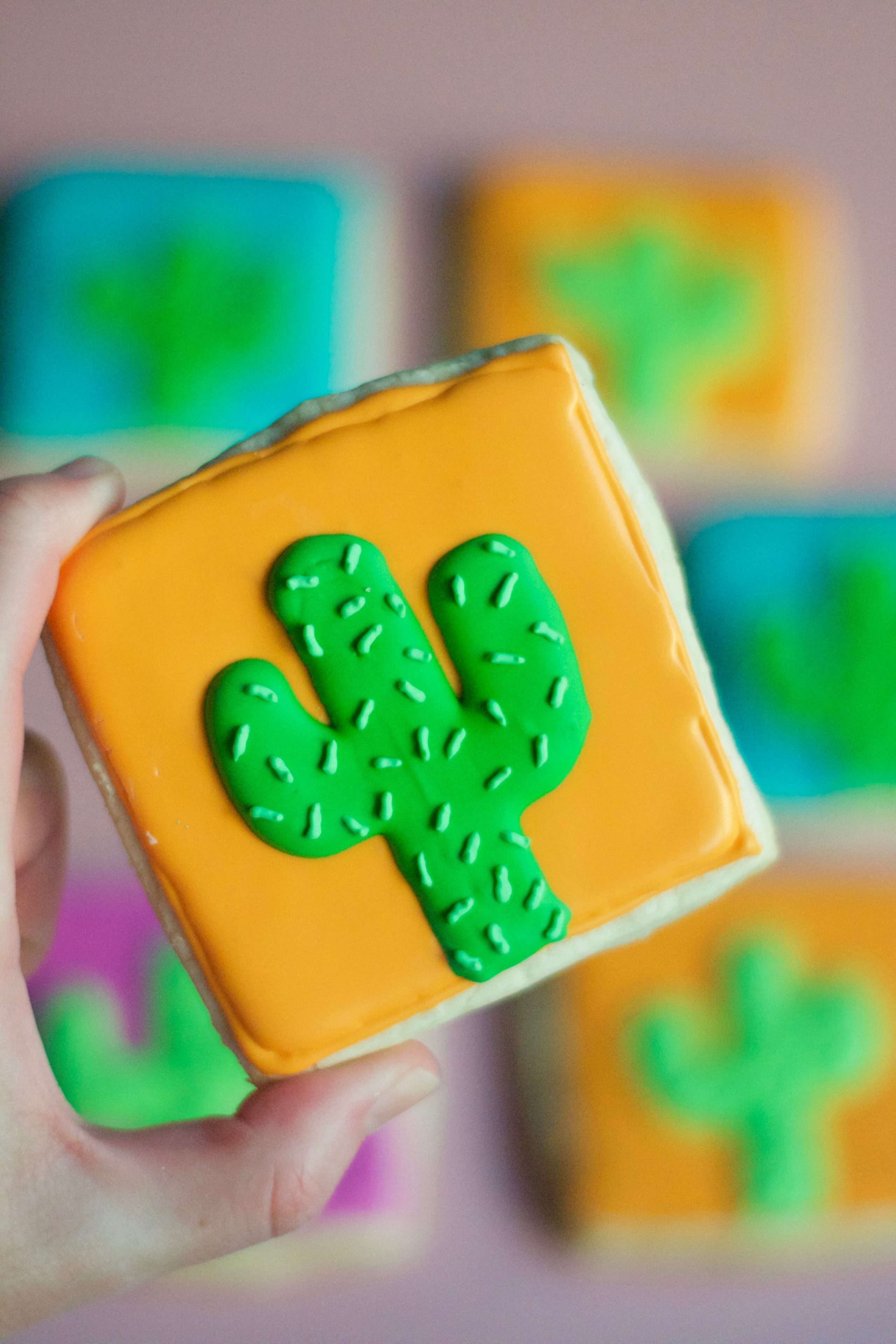 A hand holds a gluten-free cactus sugar cookie decorated with orange icing, a green cactus design, and white sprinkles, with similar colorful cactus cookies blurred in the background.