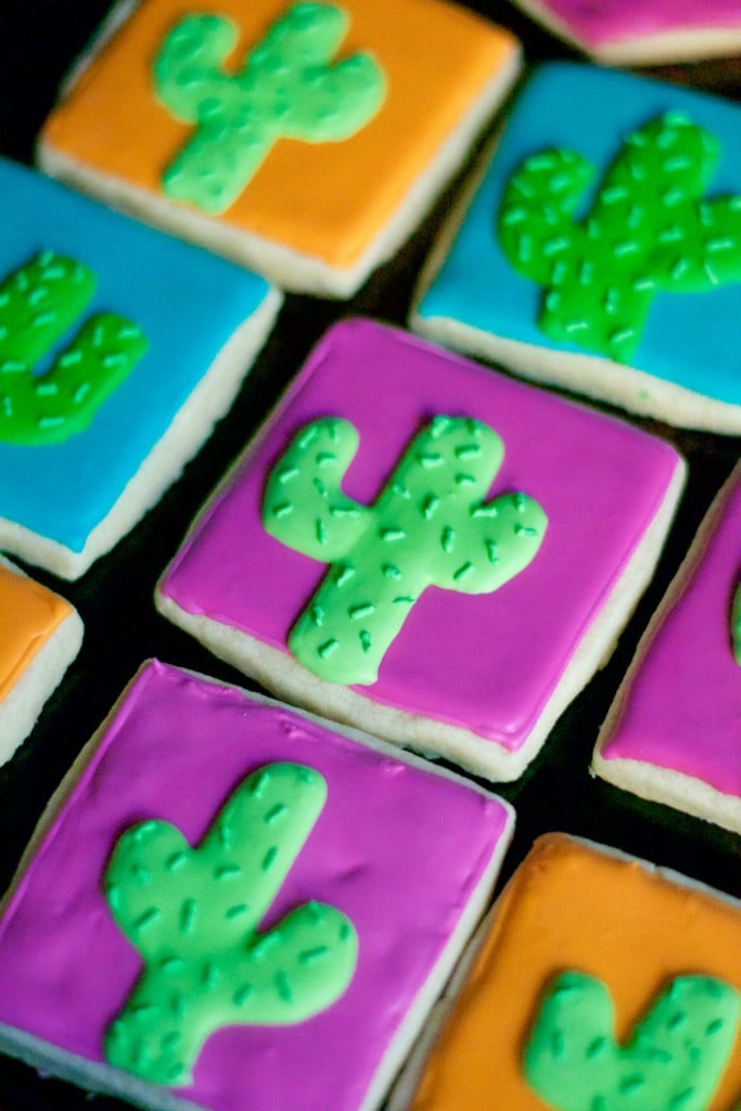 Colorful square gluten-free cactus sugar cookies decorated with green icing on bright orange, blue, and purple backgrounds. The cookies are arranged closely together on a dark surface.