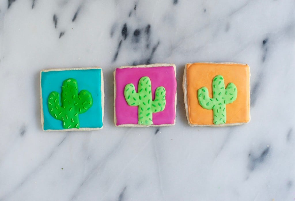 Three gluten-free cactus sugar cookies are decorated with colorful icing—one blue, one pink, and one orange—each featuring a green cactus and displayed on a marble surface.