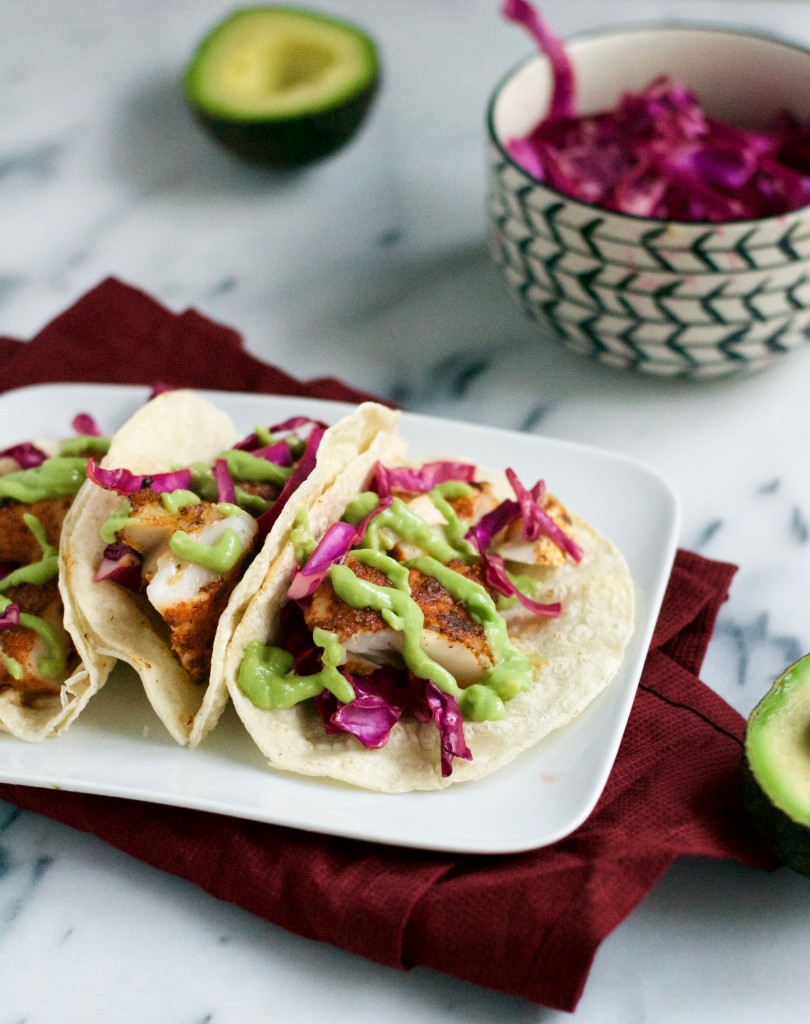 Three Blackened Fish Tacos on a white plate, topped with shredded purple cabbage and green sauce. The plate sits on a red cloth. In the background, there’s a bowl of cabbage and halved avocado on a marble surface.