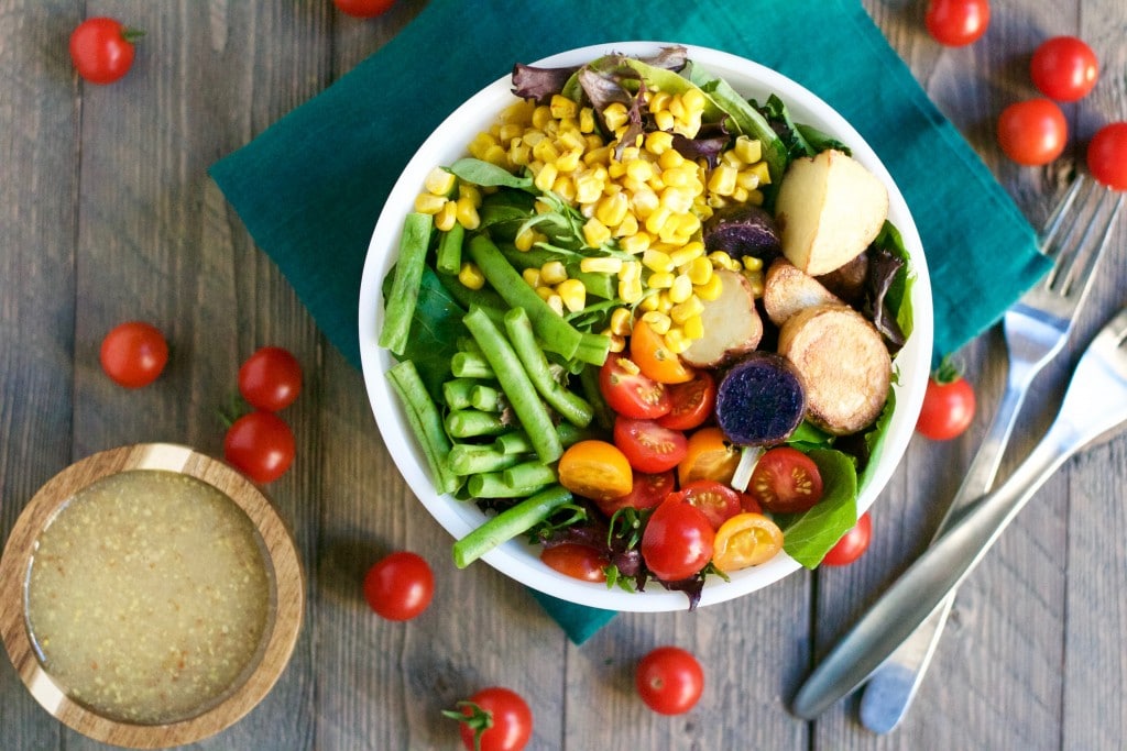 A Farmer's Market Salad with Mustard Vinaigrette sits on a green napkin, filled with cherry tomatoes, corn, green beans, potatoes, and leafy greens. Cherry tomatoes are scattered on the wooden table beside a fork, knife, and bowl of dressing.