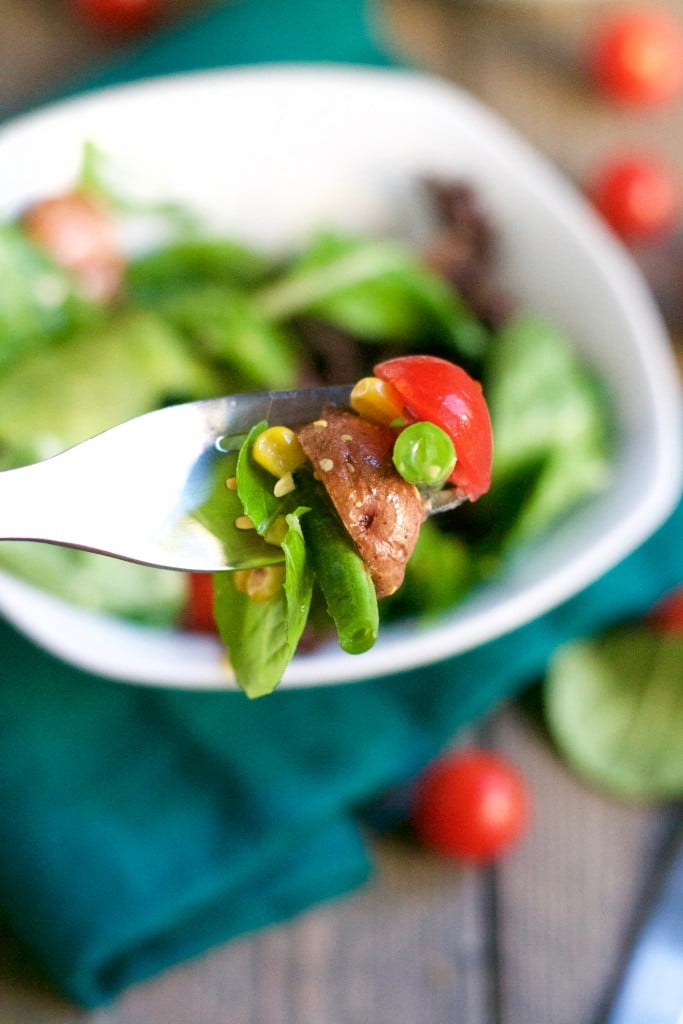A close-up of a fork holding a bite of Farmer's Market Salad with Mustard Vinaigrette—lettuce, green beans, corn, peas, tomato, and grilled chicken—with a bowl of salad blurred in the background.