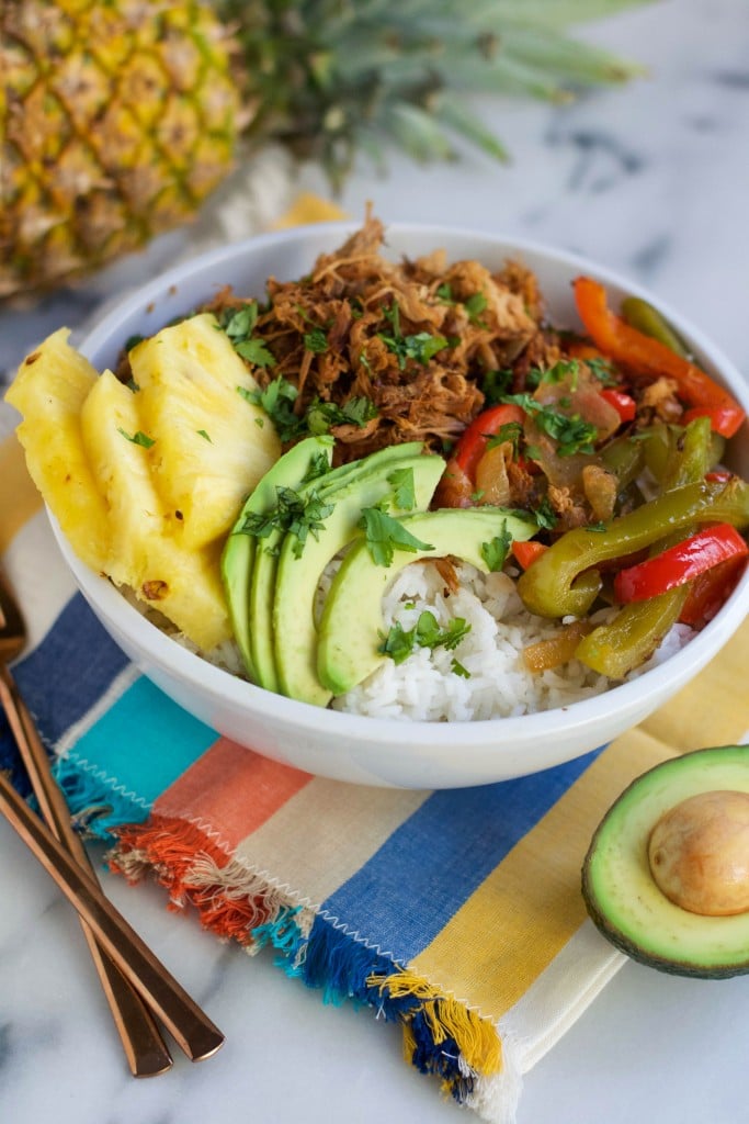 A Crock Pot Pineapple Pork Burrito Bowl filled with white rice, sliced avocado, pineapple, shredded cooked pork, and sautéed red and green bell peppers, garnished with chopped herbs. A colorful napkin and gold utensils are beside the bowl.