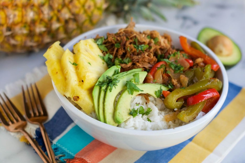 A Crock Pot Pineapple Pork Burrito Bowl features white rice topped with sliced avocado, grilled pineapple, shredded pork, sautéed red and green bell peppers, and fresh herbs, served on a striped napkin with two forks nearby.