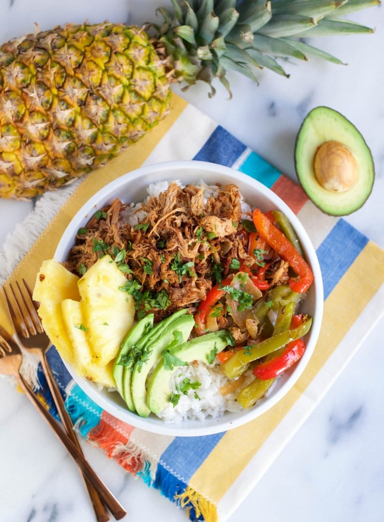 A Crock Pot Pineapple Pork Burrito Bowl features rice topped with shredded meat, sautéed bell peppers, avocado slices, and fresh pineapple, with a whole pineapple and halved avocado beside the bowl on a colorful napkin.