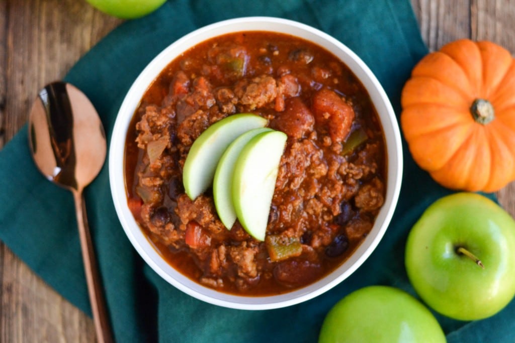 An aerial view of a bowl of pumpkin turkey chili with sliced apples on top with mini pumpkins and apples next to it.