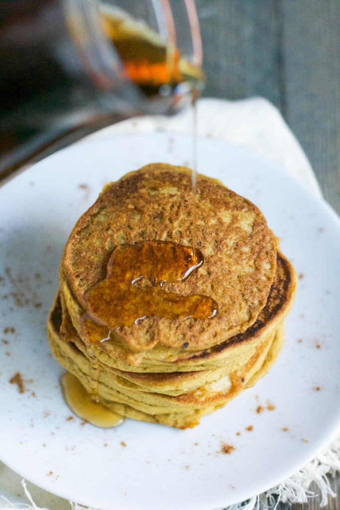 A carafe of maple syrup being drizzled over a stack of gluten-free pumpkin pancakes.