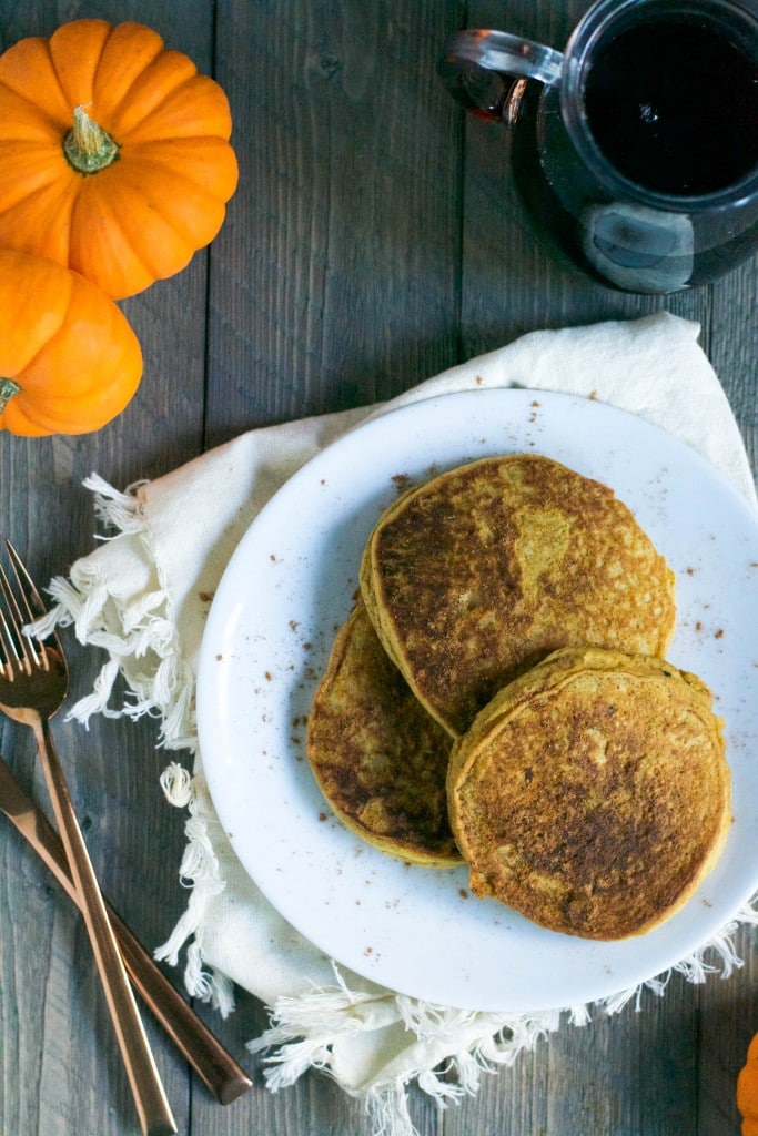 An overhead view of a plate of three pumpkin panckes with mini pumpkins on the table and a carafe of maple syrup next to it.