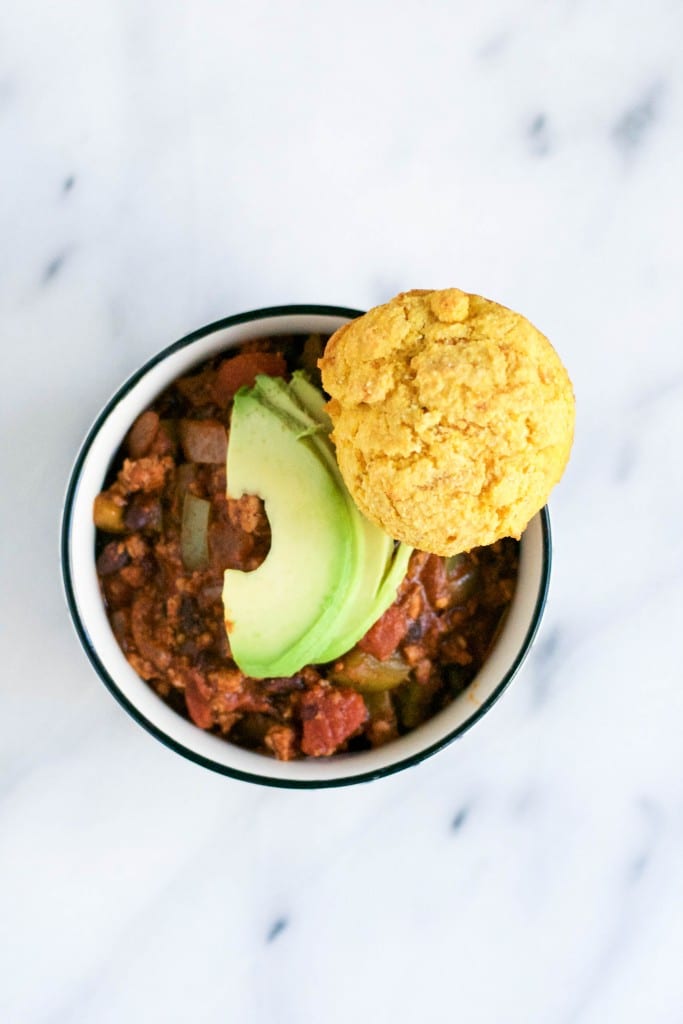 A bowl of chili topped with sliced avocado and a sweet potato cornbread muffin.