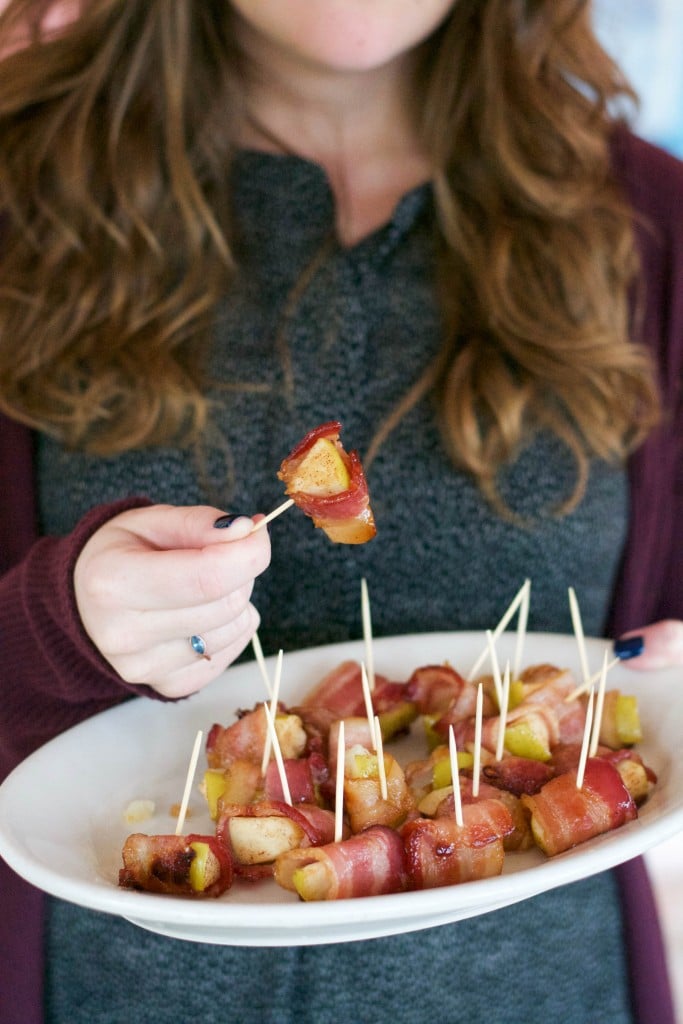 A woman holding a platter of bacon wrapped apples in one hand and picking up a piece with the other.