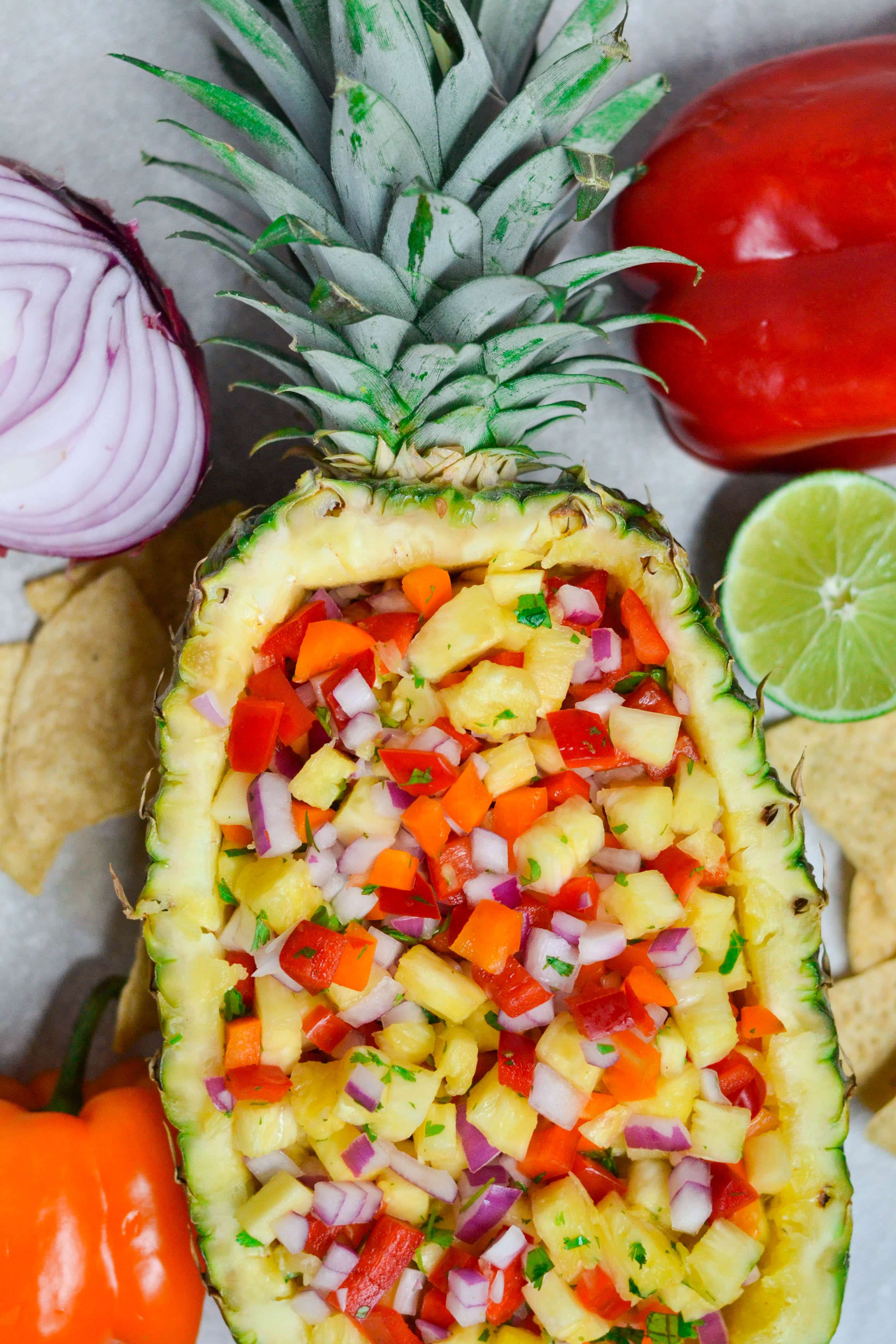 A pineapple boat filled with pineapple salsa with limes, bell peppers, tortilla chips and red onion on the table.