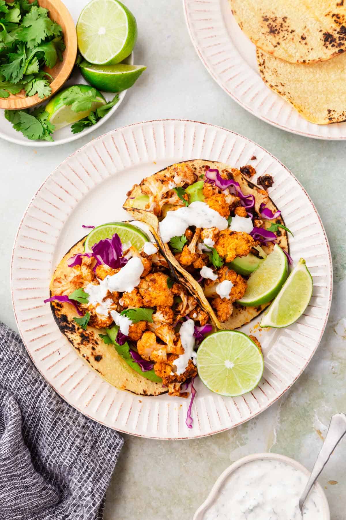 An overhead view of two cauliflower tacos on corn tortillas topped with cilantro, shredded purple cabbage, sliced avocado, cilantro and lime wedges.