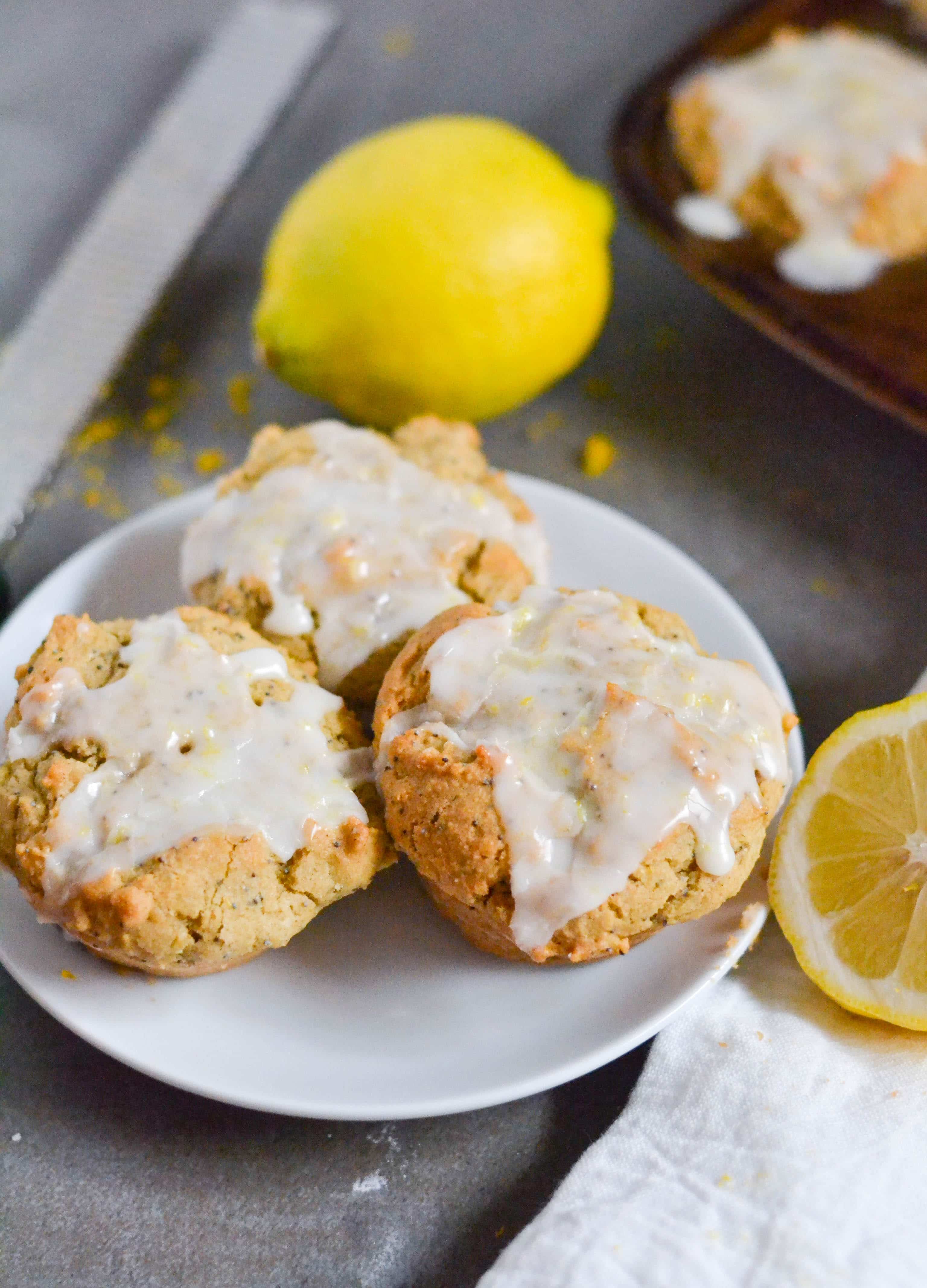 A white plate with three lemon poppyseed muffins with lemon glaze on It with lemons surrounding it.