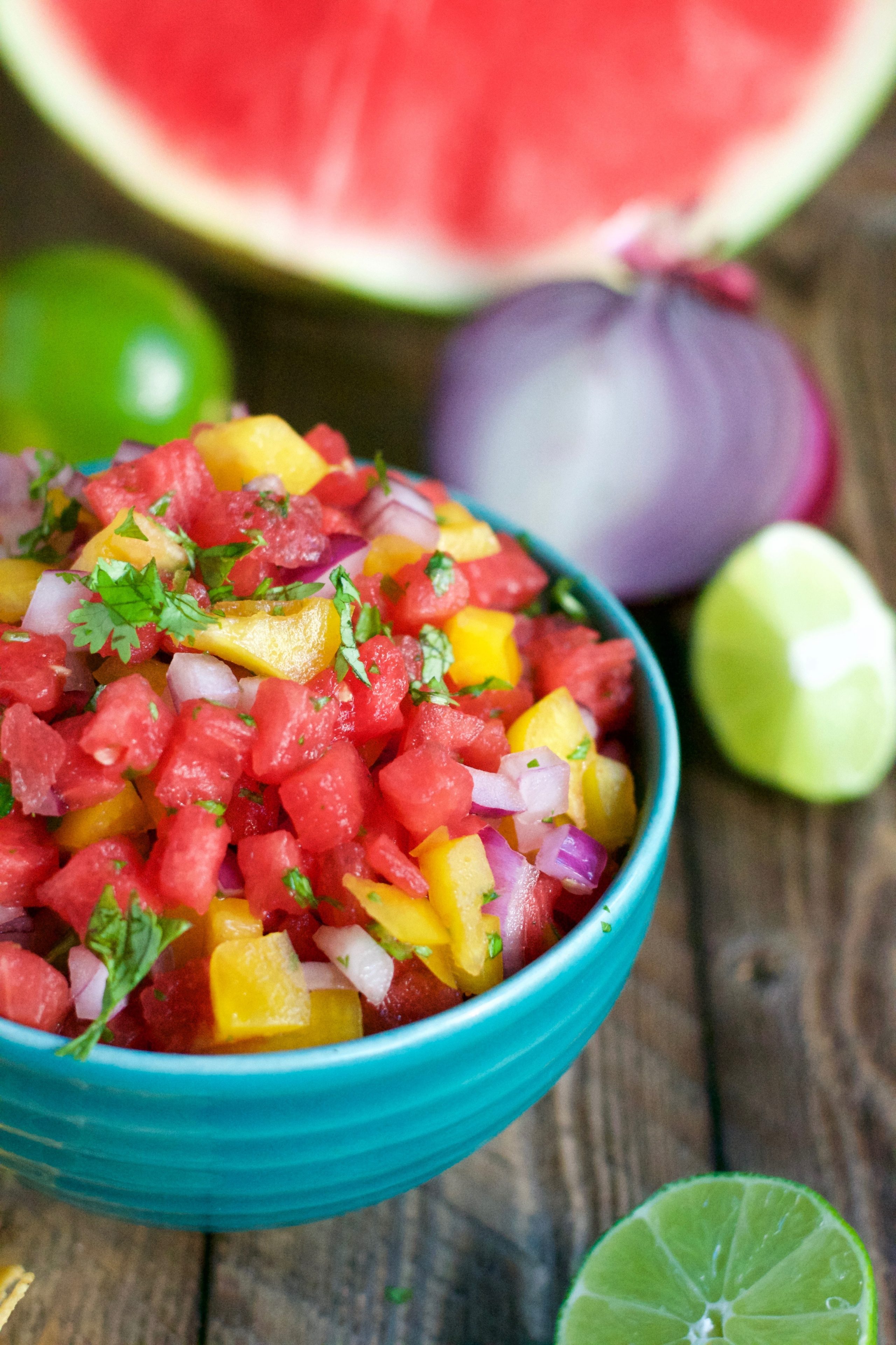 A turquoise bowl filled with vibrant watermelon salsa—diced watermelon, yellow mango, red onion, and cilantro—sits on a wooden table, surrounded by lime halves and a slice of watermelon.