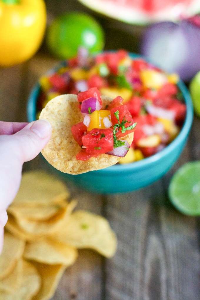 A hand holds a tortilla chip topped with vibrant watermelon salsa, featuring diced tomatoes, yellow peppers, red onion, and cilantro. A blue bowl of salsa and more chips are visible in the background on a wooden surface.