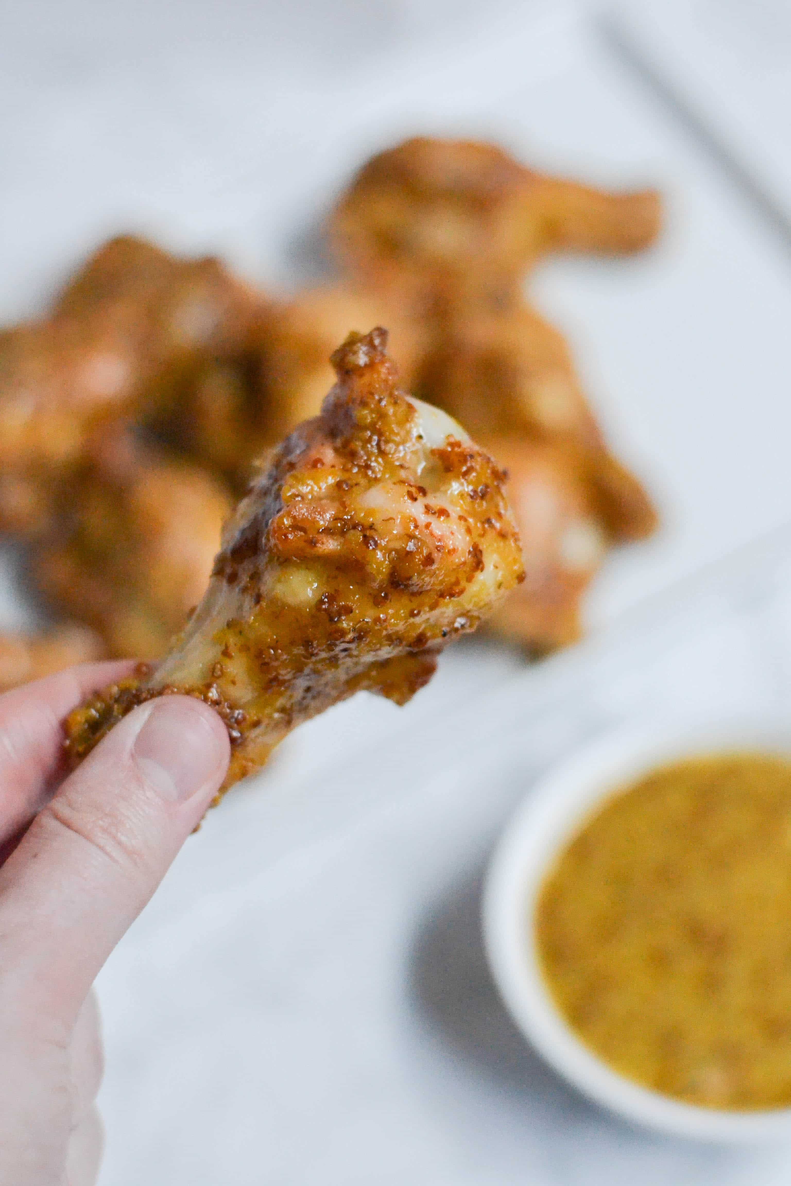 A hand holds a saucy Honey Mustard Baked Chicken Wing, with more wings blurred in the background and a small bowl of mustard dipping sauce nearby.