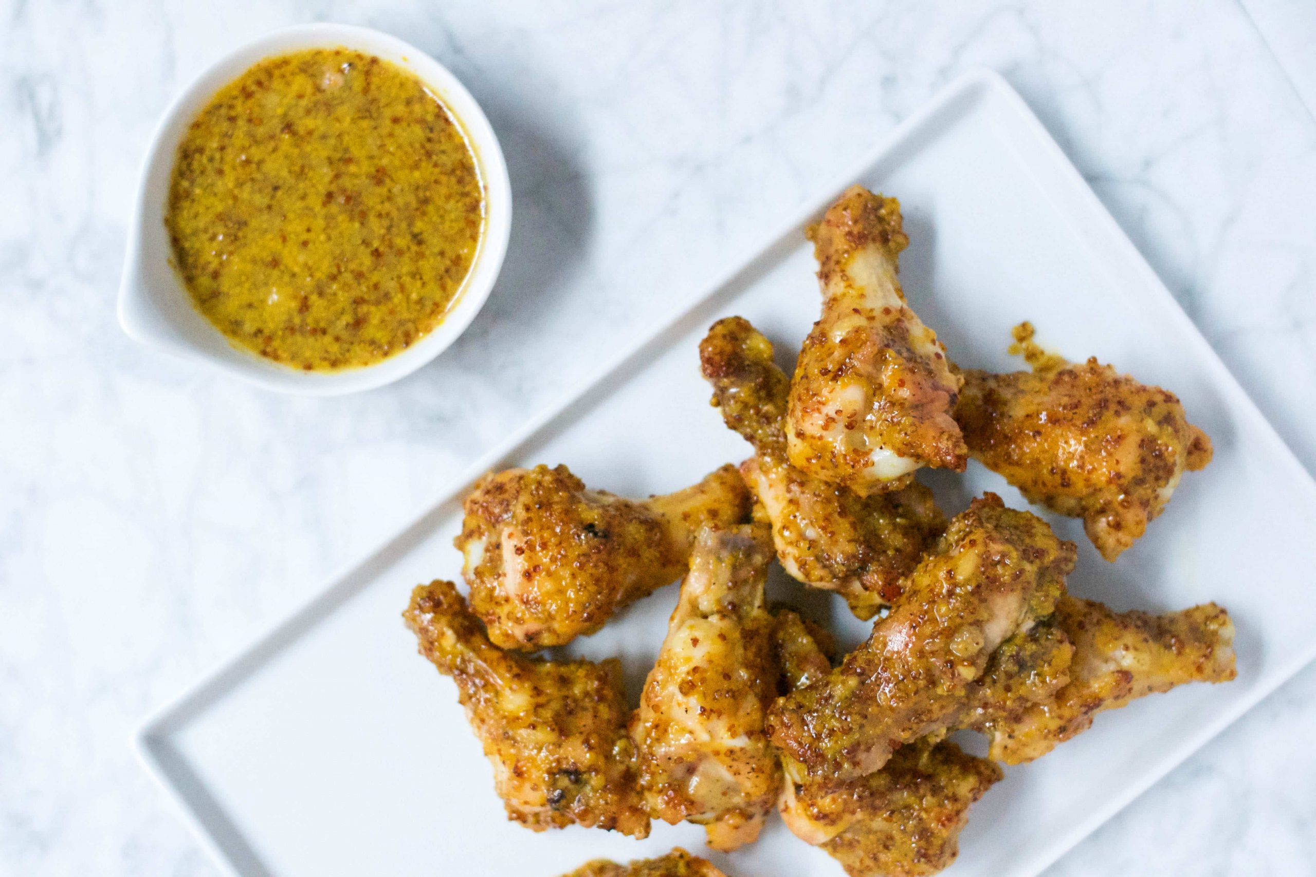 A white rectangular plate holds several pieces of crispy, Honey Mustard Baked Chicken Wings. Beside the plate, a small white bowl brims with grainy mustard dipping sauce, all set on a bright white marble surface.