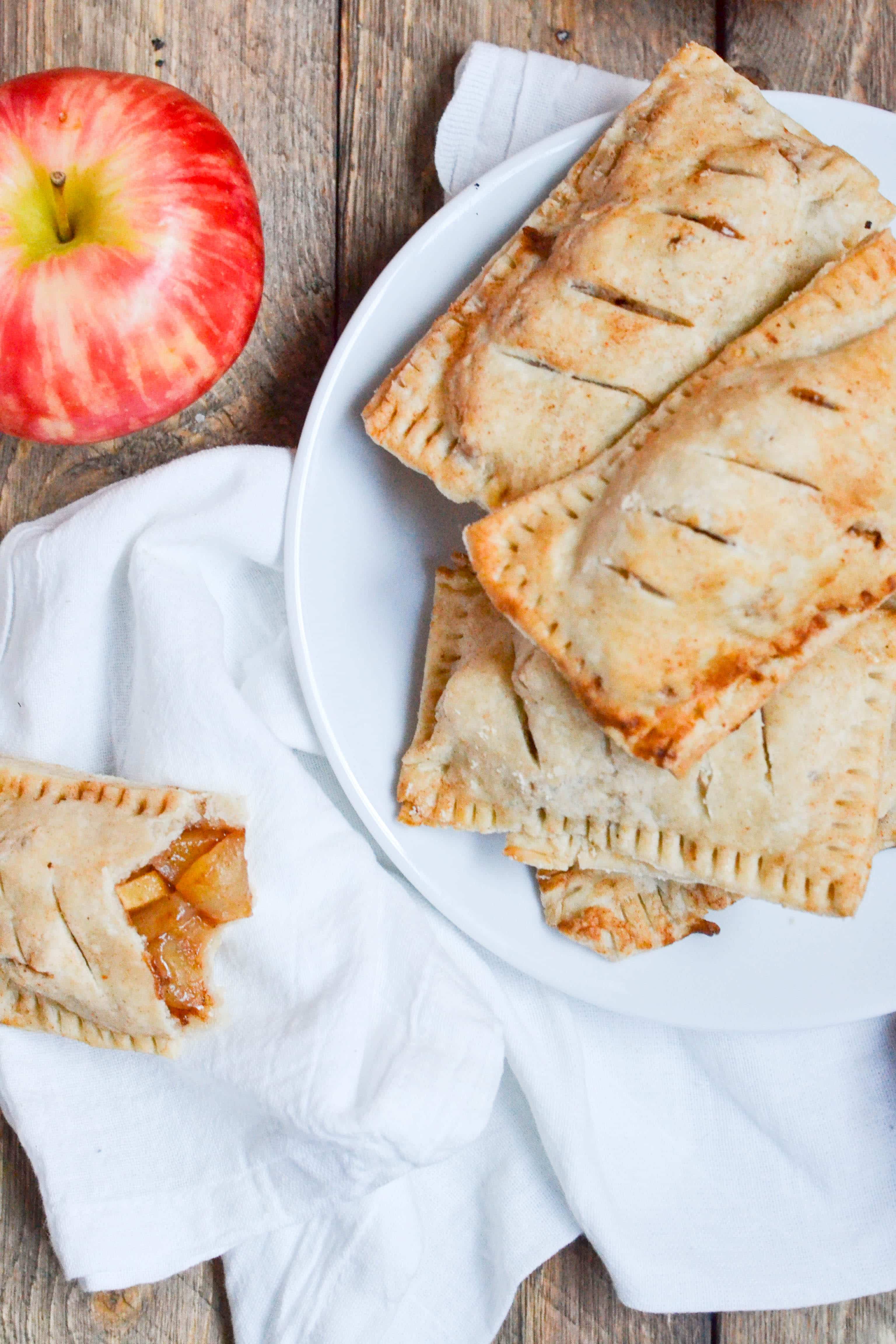 A white plate with a stack of apple hand pies on them with a fresh apple next to it.