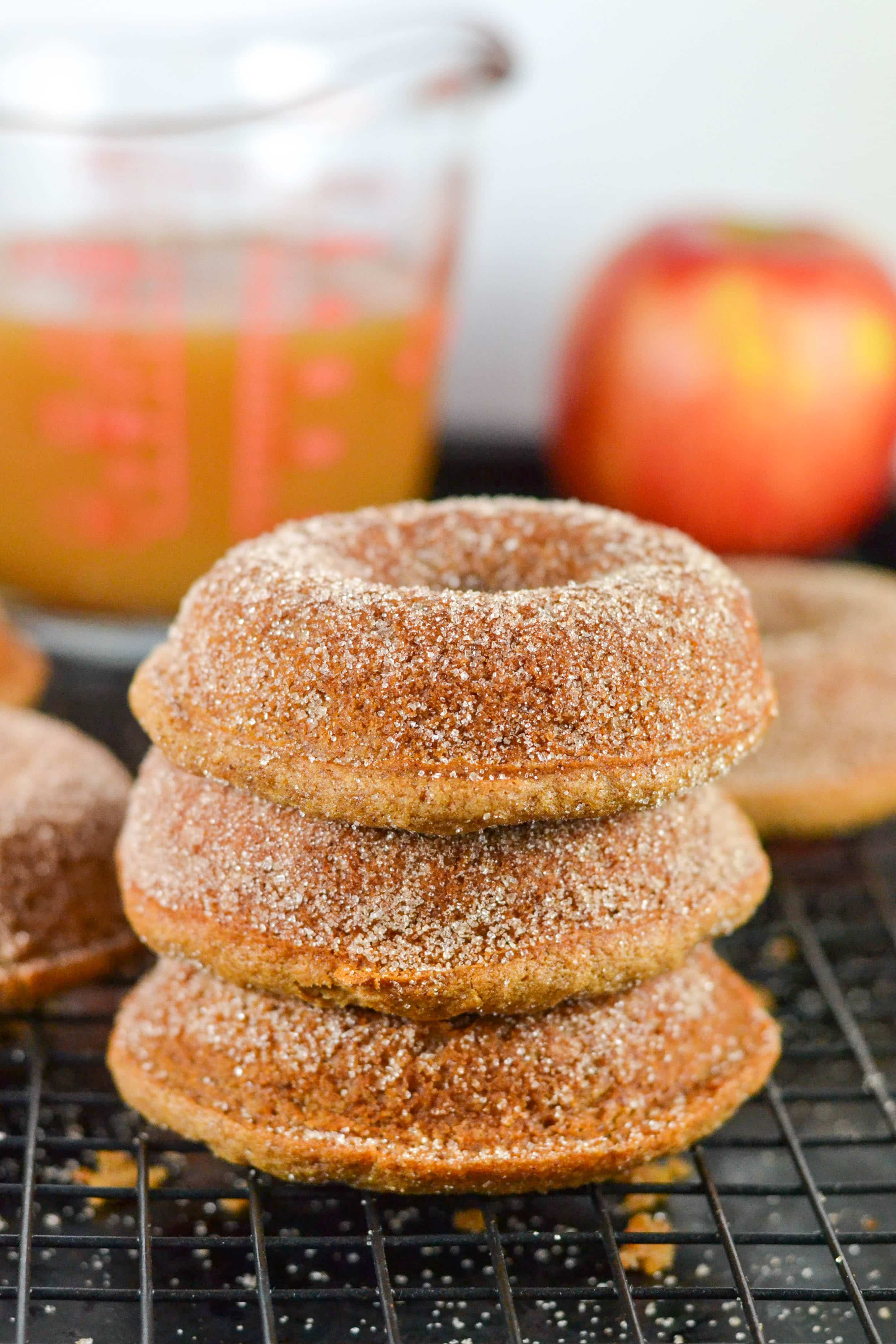 Four cinnamon sugar-coated gluten-free vegan apple cider donuts are stacked on a cooling rack, with a red apple and a glass measuring cup filled with brown liquid in the blurred background.