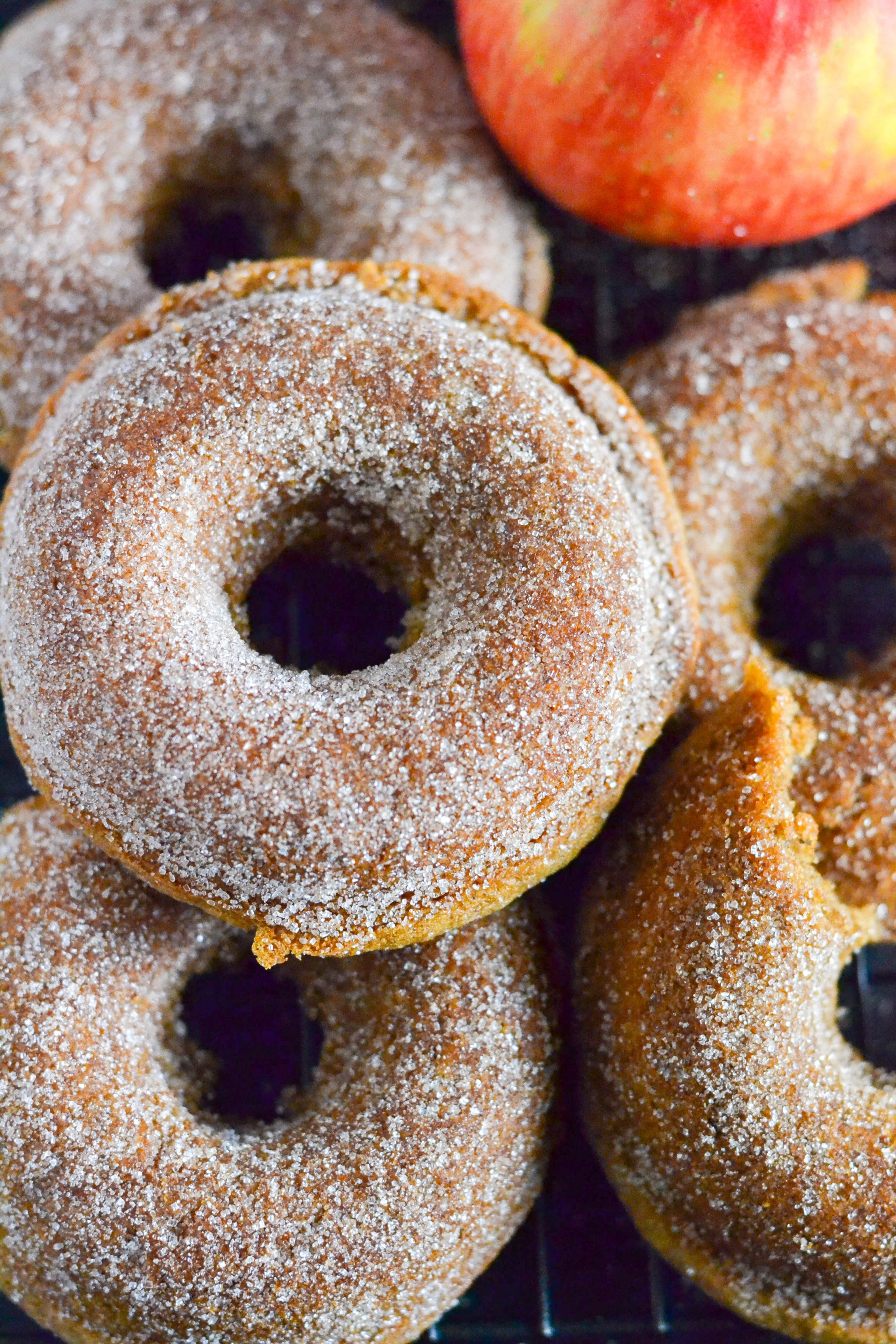Five sugar-coated gluten-free vegan apple cider donuts sit closely together, with part of a red apple visible in the corner. The donuts appear soft and are displayed on a cooling rack.