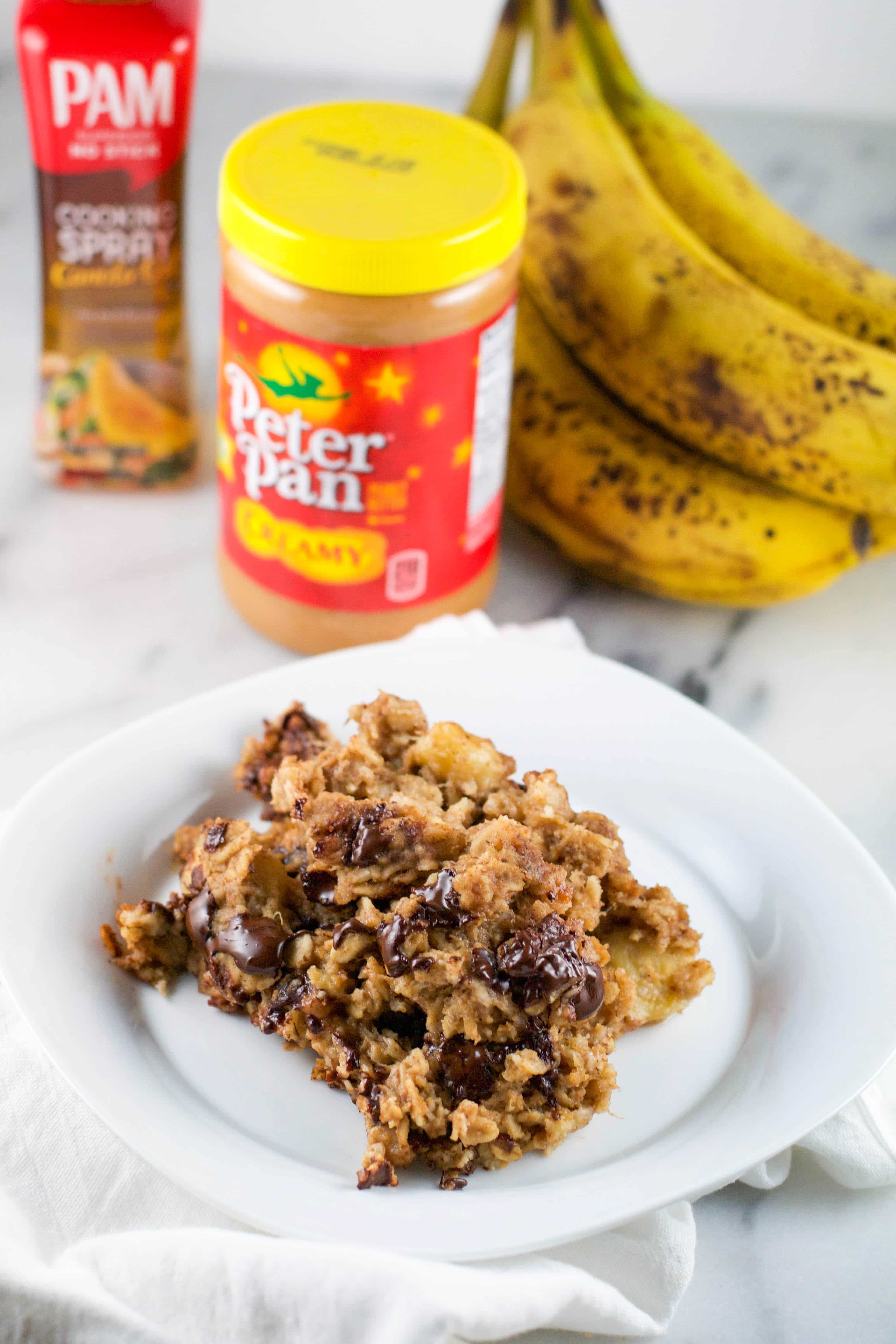 A white plate with chocolate peanut butter banana baked oatmeal with peanut butter, cooking spray and bananas in the background.