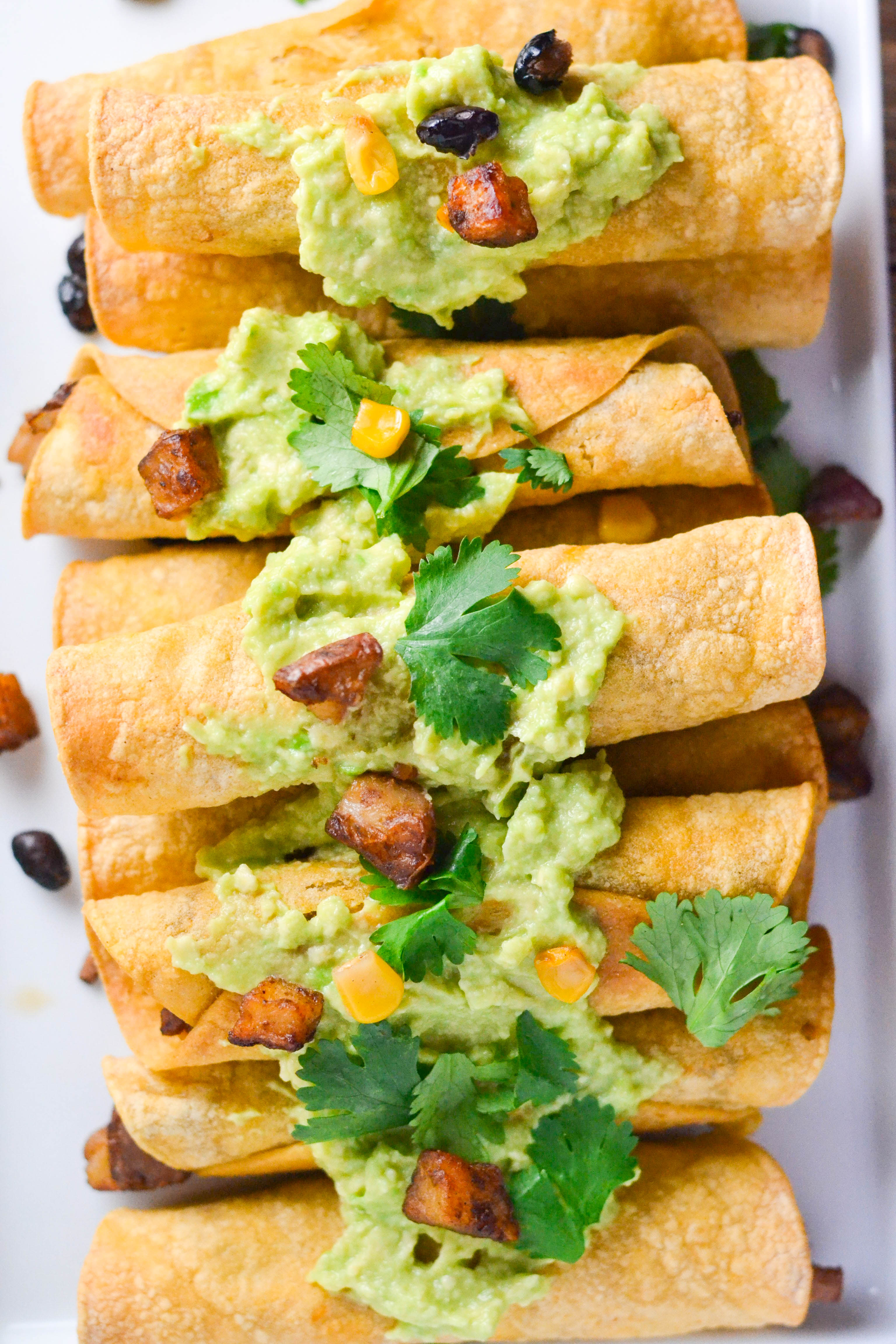 A platted filled with stacks of Red Potato and Black Bean Taquitos topped with avocado, corn, and cilantro.