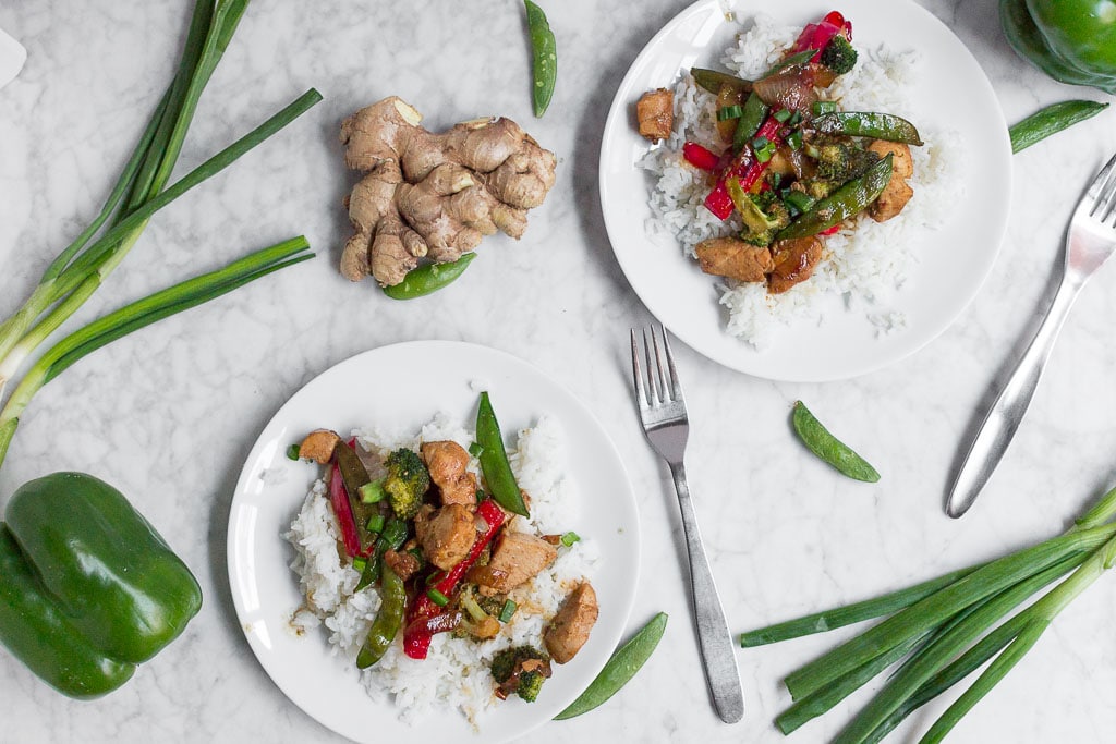 Two white plates with rice topped with Chicken Teriyaki Stir Fry—juicy chicken, broccoli, snap peas, and red bell peppers. Surrounding the plates are fresh ginger, green onions, a green bell pepper, and snap peas on a marble surface.