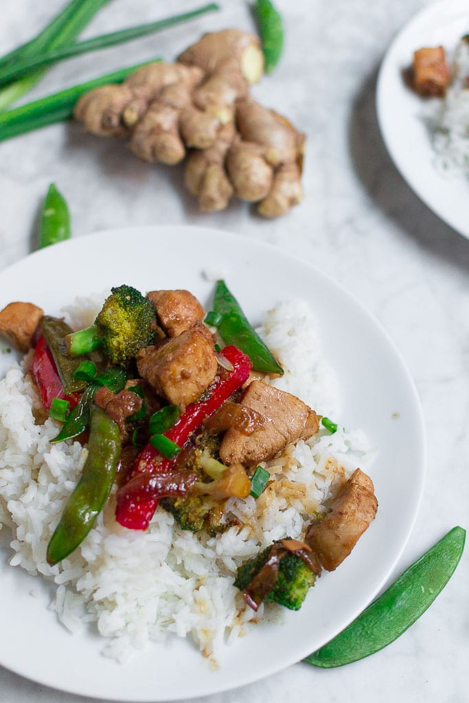 A white plate with white rice topped with Chicken Teriyaki Stir Fry, featuring stir-fried chicken, broccoli, snap peas, and red bell pepper. In the background are fresh ginger, green onions, and another plate partially visible.
