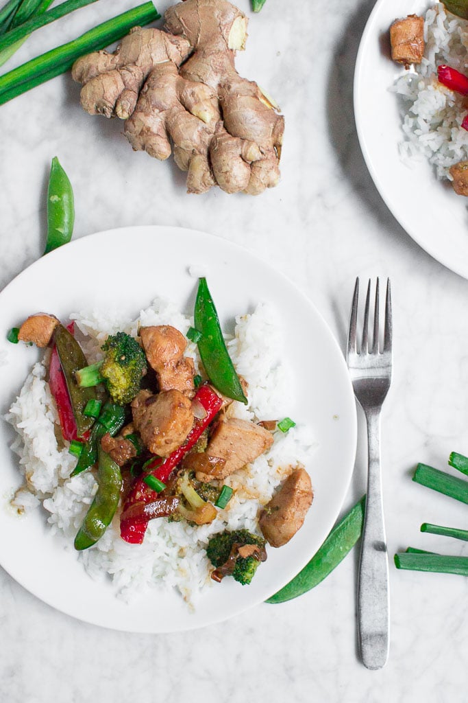 A white plate with rice topped with Chicken Teriyaki Stir Fry—featuring chicken, broccoli, snap peas, and red bell peppers—sits on a marble surface next to a fork. Fresh ginger, green onions, and snap peas are nearby.