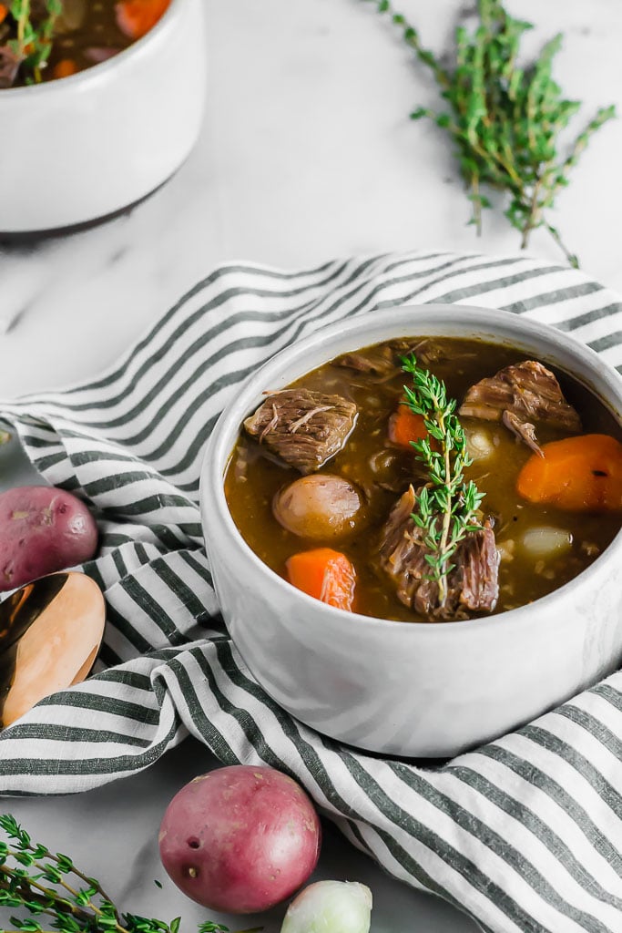 A white bowl of gluten-free beef stew with carrots, potatoes, and herbs sits on a striped cloth, garnished with a sprig of thyme. Whole red potatoes and fresh thyme are nearby on a white marble surface.