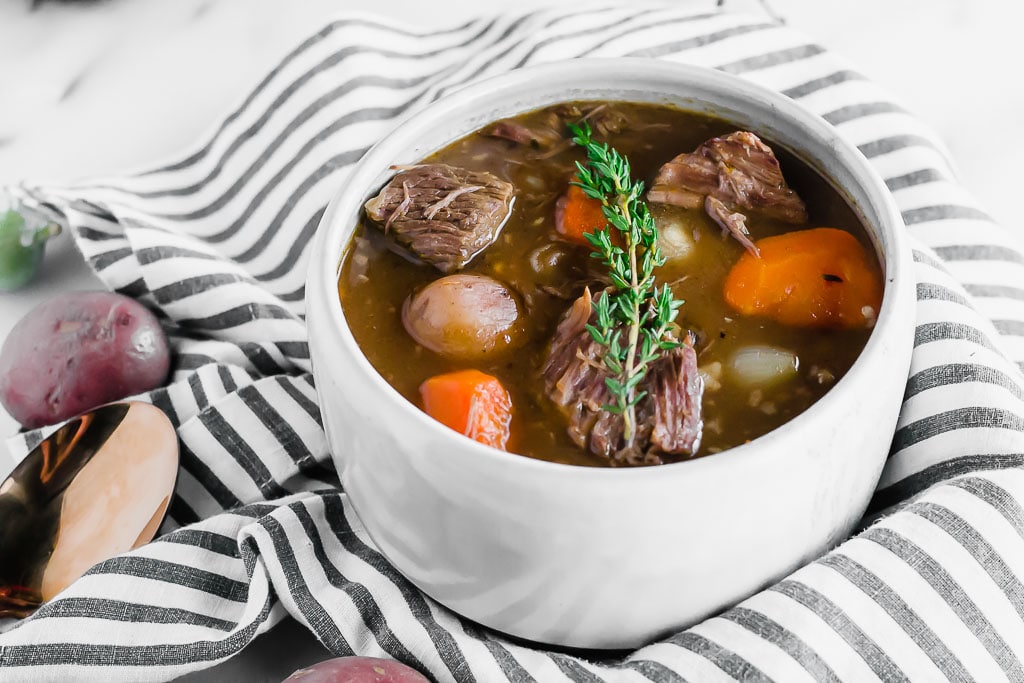 A white bowl filled with gluten-free beef stew, containing chunks of beef, carrots, potatoes, and garnished with fresh thyme, sits on a black and white striped cloth. Purple potatoes and a spoon are nearby.