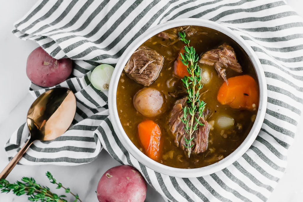 A bowl of gluten-free beef stew with chunks of beef, carrots, potatoes, and onions, garnished with fresh thyme, sits on a striped cloth alongside a gold spoon and red potatoes.