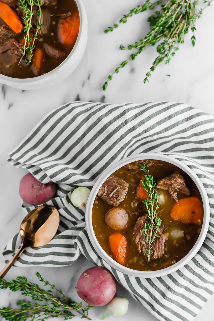 Two bowls of gluten-free beef stew with carrots, potatoes, and herbs sit on a white marble surface. One bowl rests on a striped cloth with scattered potatoes, onions, thyme, and a gold spoon nearby.